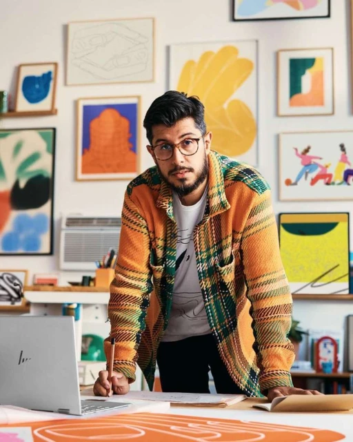 Bearded man in glasses and orange flannel stands near a laptop with colorful abstract artwork and framed prints on a white wall.