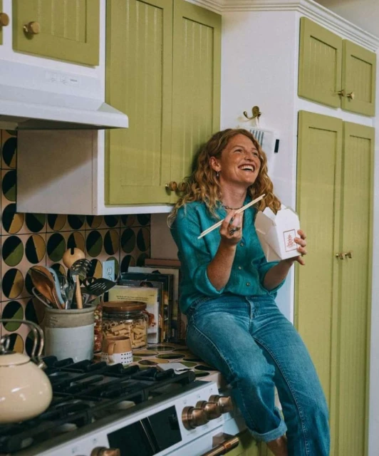 A woman with curly hair sits on a kitchen counter with a box of Chinese takeout and chopsticks, wearing jeans and a teal button-down shirt.