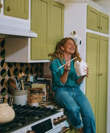 A woman with curly hair sits on a kitchen counter with a box of Chinese takeout and chopsticks, wearing jeans and a teal button-down shirt.