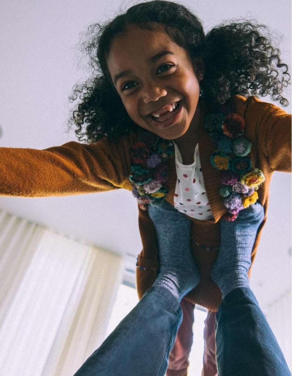 Smiling child with curly hair wearing a colorful flower-decorated cardigan being held up by a person wearing jeans indoors.