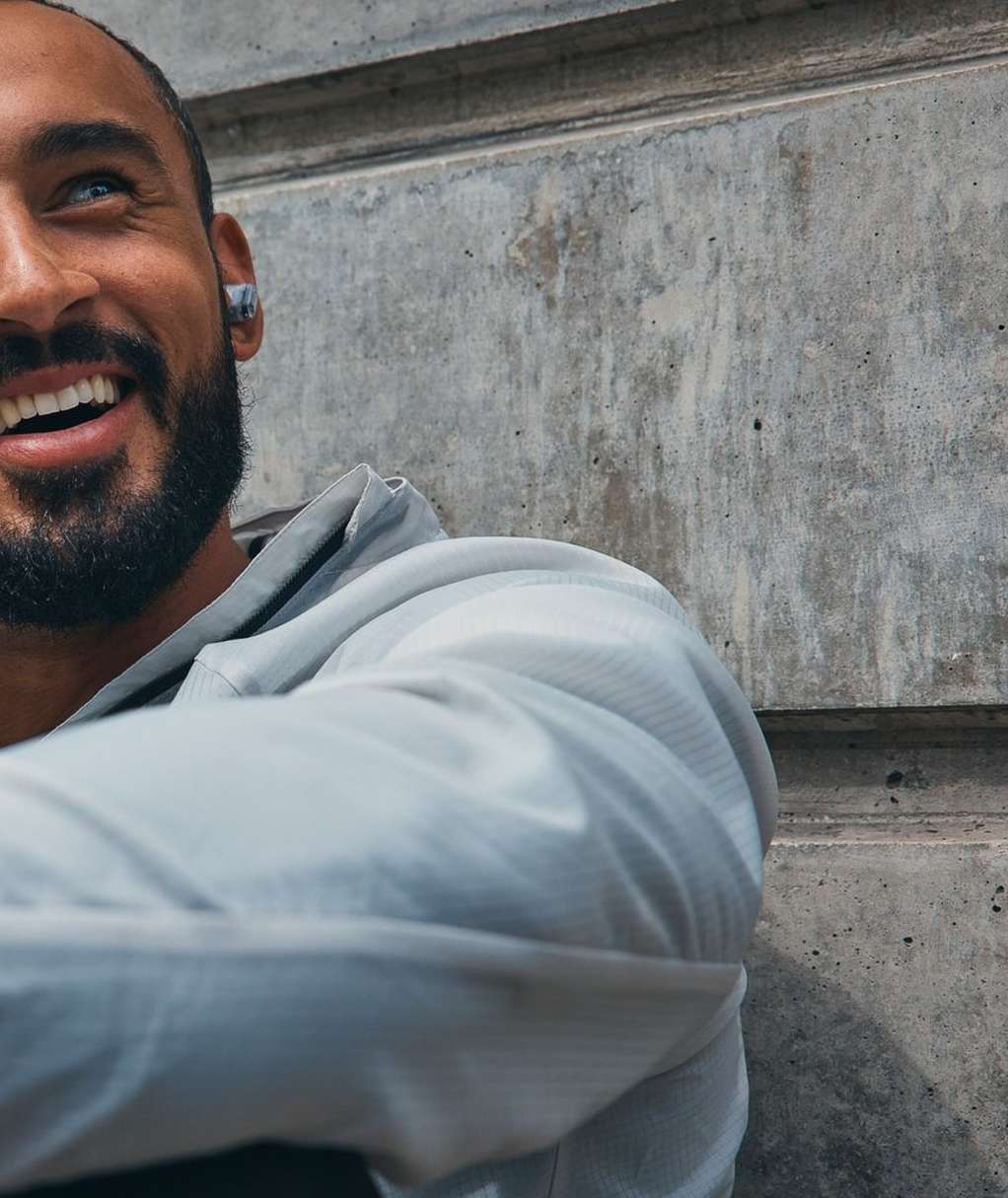 Close up shot of a happy adult male smiling while wearing wireless earbuds and a grey hoodie with a concrete wall background.