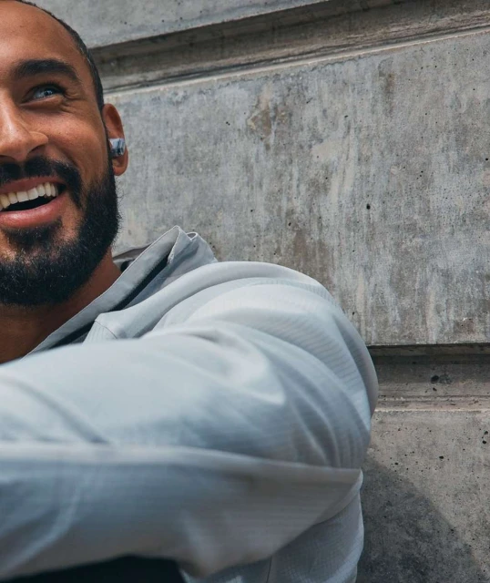 Close up shot of a happy adult male smiling while wearing wireless earbuds and a grey hoodie with a concrete wall background.