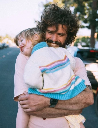 A father with curly hair holds his child close in an outdoor setting, both wearing casual clothing on a sunny day.