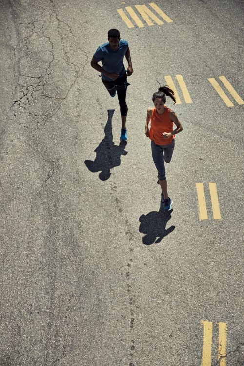 High-angle view of a man and woman running together on a cracked asphalt surface with yellow painted stripes. Shadows are visible.