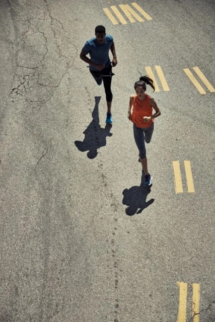 High-angle view of a man and woman running together on a cracked asphalt surface with yellow painted stripes. Shadows are visible.
