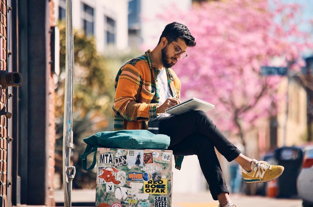 Man with glasses sitting outdoors on a box with stickers, writing on a tablet. He wears a patterned jacket and yellow sneakers.
