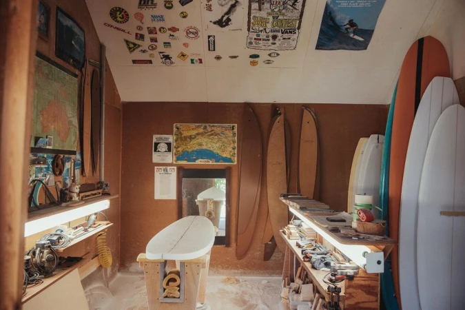 Surfboards stand in a workshop, illuminated by soft light. Various tools, maps, and vintage surf posters adorn the walls. 