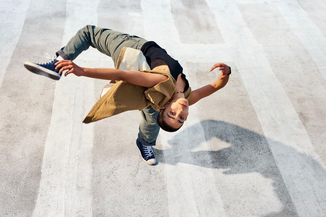 A young woman in casual clothing performs an acrobatic move outdoors on a concrete surface with white stripes, captured from a high angle.