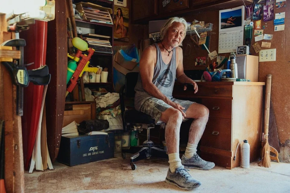 Man with long gray hair sits in an office chair at a desk cluttered with surfing memorabilia, calendars, and water guns inside a garage.