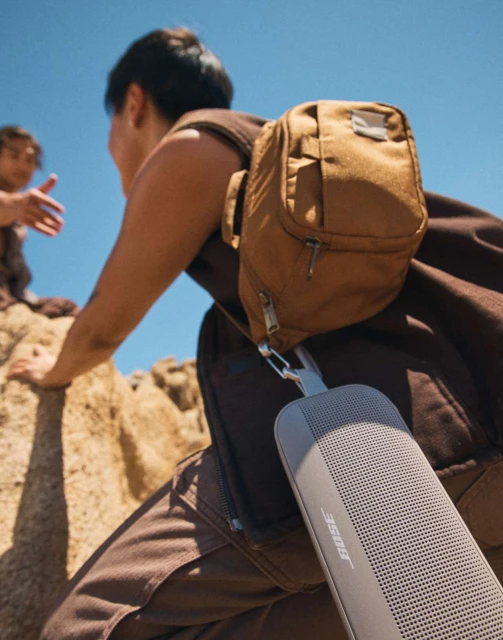 A person wearing a backpack and a portable speaker climbing rocks on a sunny day with clear blue skies, and another person in the background.