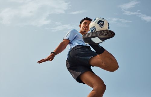 A low-angle shot of an adult male athlete balancing a soccer ball on his foot against a bright, cloudy sky.