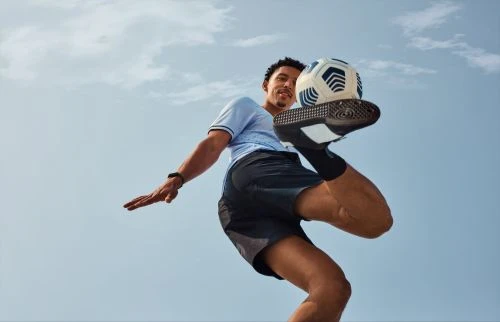 A low-angle shot of an adult male athlete balancing a soccer ball on his foot against a bright, cloudy sky.