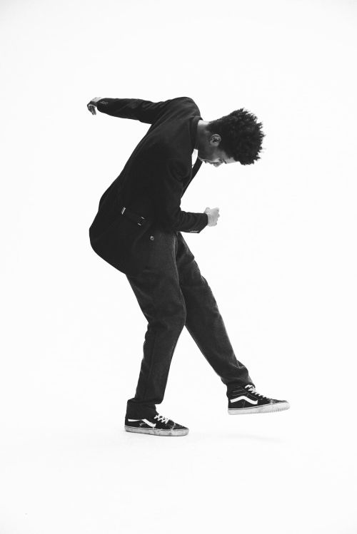 A Black male dancer dressed in black suit and sneakers strikes a dynamic pose against a stark white backdrop, creating a striking monochrome image.