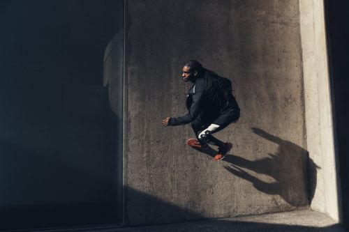 Athletic Black man leaps in urban setting, wearing dark athletic wear and red shoes, illuminated by sunlight against a concrete wall.