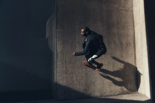 Athletic Black man leaps in urban setting, wearing dark athletic wear and red shoes, illuminated by sunlight against a concrete wall.