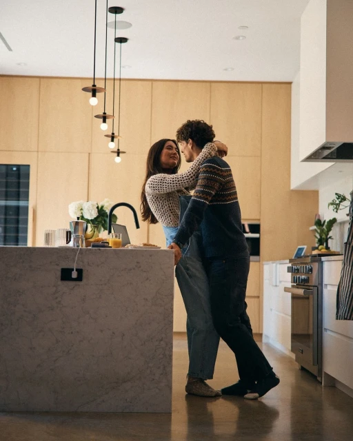 A young couple embraces in a naturally-lit kitchen. The woman faces the camera while the man is in profile. Kitchen island and appliances visible.