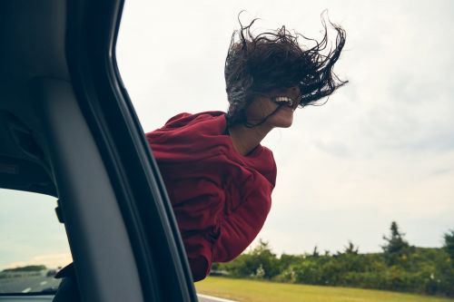 Woman in red leans out of a car window with blowing dark hair, smiling with trees and sky visible beyond the vehicle and roadside.