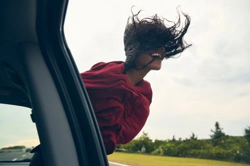 Woman in red leans out of a car window with blowing dark hair, smiling with trees and sky visible beyond the vehicle and roadside.