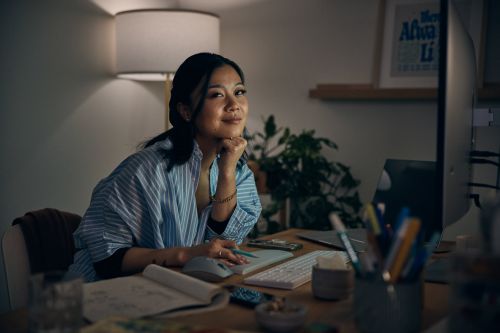 Smiling woman works at her desk with a computer, notebook, pens, and lighting in a cozy interior setting at night.