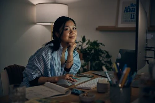 Smiling woman works at her desk with a computer, notebook, pens, and lighting in a cozy interior setting at night.