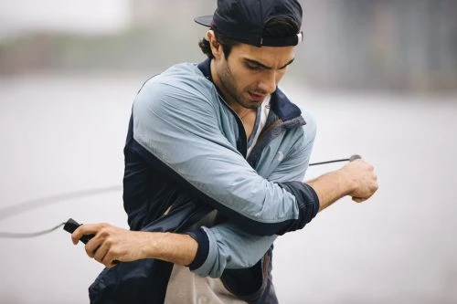 Adult male wearing a baseball cap exercises outdoors with a jump rope, wearing a light blue jacket, with a blurred background.