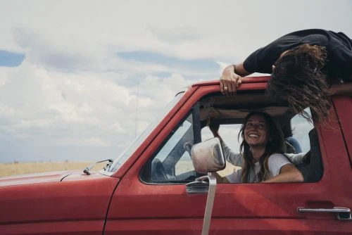 Two young women enjoy a road trip in a red pickup truck, with one sitting in the driver's seat and the other half outside the truck.