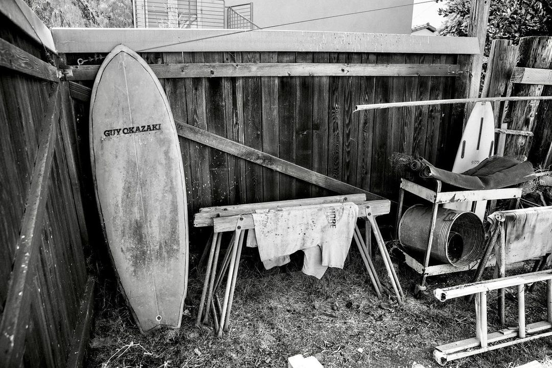 Monochrome image features a surfboard leaning against a wooden fence, with a sawhorse, metal bucket, and various items nearby.