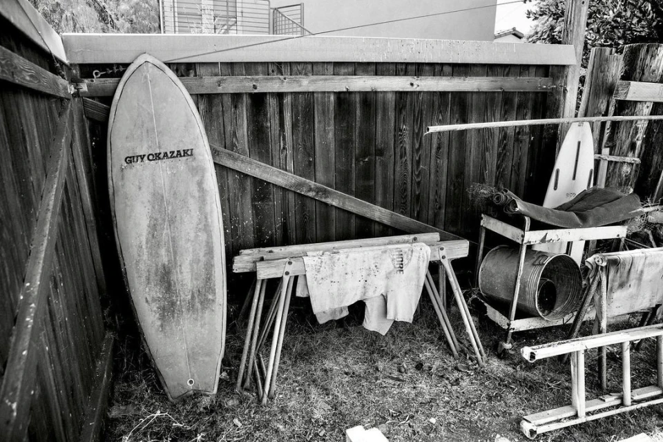 Monochrome image features a surfboard leaning against a wooden fence, with a sawhorse, metal bucket, and various items nearby.