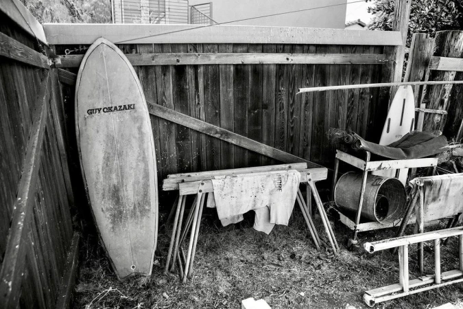 Monochrome image features a surfboard leaning against a wooden fence, with a sawhorse, metal bucket, and various items nearby.