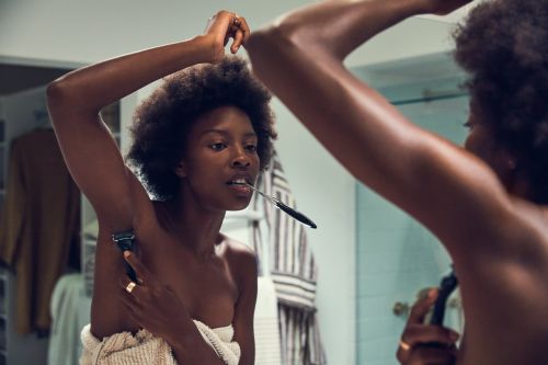Woman in bathroom shaving armpit with razor. Dark-skinned adult female with afro hair, towel wrapped around body, and bathroom setting.