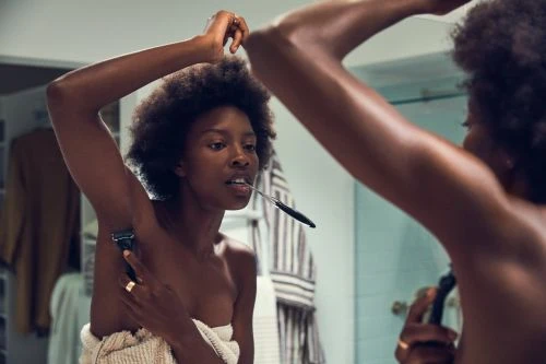 Woman in bathroom shaving armpit with razor. Dark-skinned adult female with afro hair, towel wrapped around body, and bathroom setting.