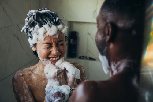 Laughing couple in shower covered in soap suds; adult woman and man; indoor setting with marble tile.