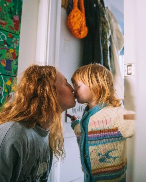 A heartwarming moment captures a mother with long red hair sharing a kiss with her young child in a cozy indoor environment.