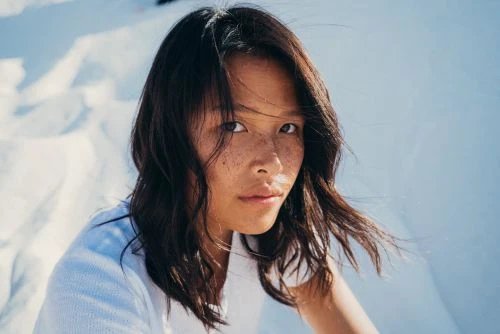 Close-up portrait of a young woman with dark hair and freckles wearing a white top against a bright sandy backdrop.
