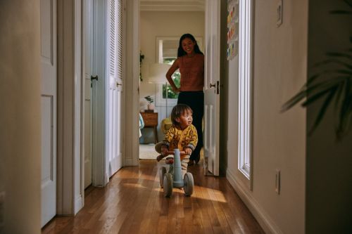 A toddler rides a blue toy tricycle down a wooden floor hallway toward the camera as an adult woman watches from a doorway in the background.