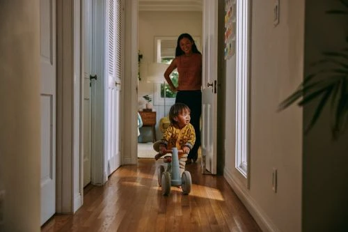 A toddler rides a blue toy tricycle down a wooden floor hallway toward the camera as an adult woman watches from a doorway in the background.