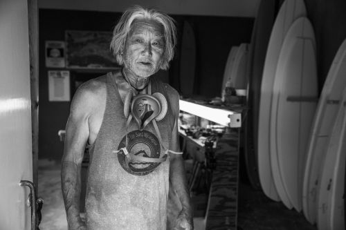 Monochrome portrait of an elderly man wearing a tank top and pendant, standing indoors near surfboards and equipment in a workshop.