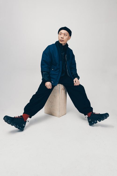 Adult male in a dark blue jacket and black pants sitting on a tan square block wearing sneakers in a studio setting.