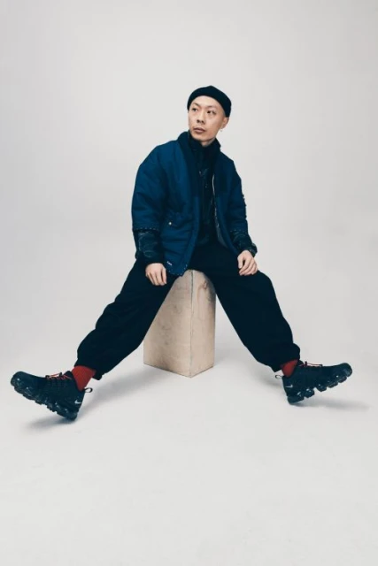Adult male in a dark blue jacket and black pants sitting on a tan square block wearing sneakers in a studio setting.