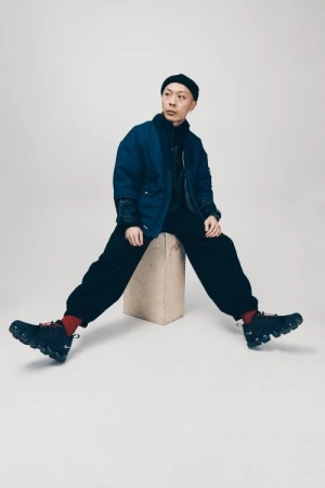 Adult male in a dark blue jacket and black pants sitting on a tan square block wearing sneakers in a studio setting.