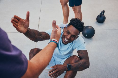 Smiling African-American man high-fives a friend after workout in a bright gym. Features fitness equipment and gray floor.