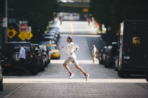 Man running across a city street in athletic wear, with parked cars and buildings visible in the background on a sunny day.