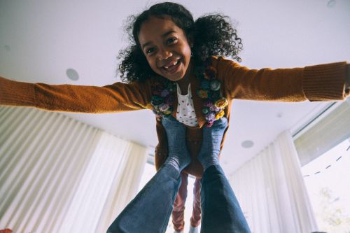 Smiling child is held aloft by adult wearing jeans and socks in a bright, modern indoor space with neutral tones and lots of natural light.