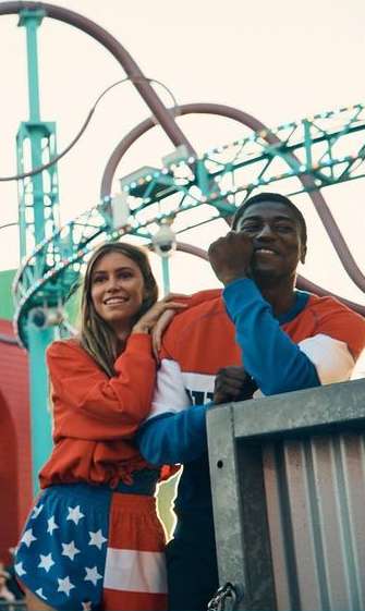 A smiling couple in matching patriotic-themed outfits stands in front of a teal and purple roller coaster at an amusement park.