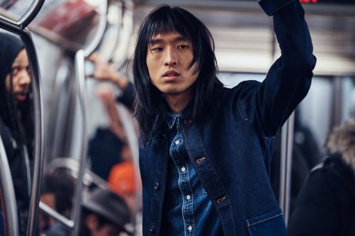 Asian man with long hair on subway wearing a denim jacket and button-down shirt, looking straight ahead with passengers in background.