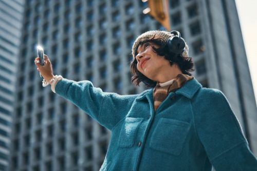 Young woman takes a selfie in an urban setting, wearing a teal jacket, knit hat, and headphones, with tall buildings as a backdrop.