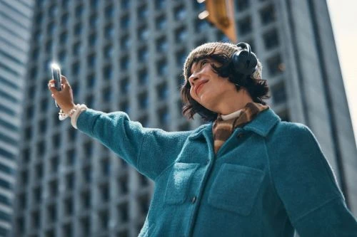 Young woman takes a selfie in an urban setting, wearing a teal jacket, knit hat, and headphones, with tall buildings as a backdrop.