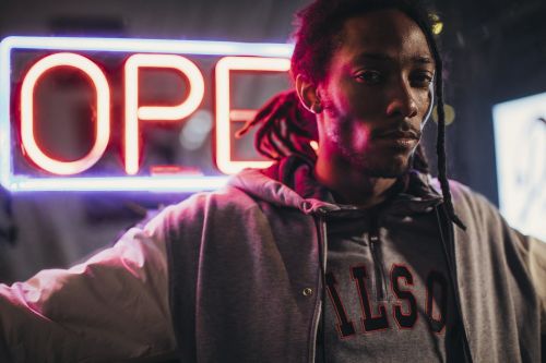 Low angle studio shot of an adult male wearing a grey hooded sweatshirt under a light jacket in front of a neon sign.