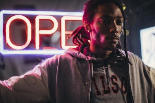 Low angle studio shot of an adult male wearing a grey hooded sweatshirt under a light jacket in front of a neon sign.