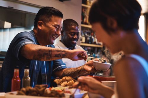 Three adults, two men and one woman, sit at a bar enjoying food and drinks in a warmly lit restaurant setting, sharing a laugh together.